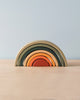 A stack of Handmade Mini Rainbow Stacker - Jungle toy pieces in gradient shades from dark green to red, arranged in an arch shape on a wooden surface against a plain light blue background.