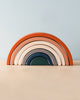 A Handmade Rainbow Stacker - Terracotta consisting of nested arches in shades of red, orange, pink, and teal arranged in size order on a wooden surface against a pale blue background.