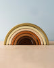 A Handmade Rainbow Stacker - Olive with seven arches, each painted in shades of brown, beige, and mustard yellow using non-toxic paint, is displayed on a light wooden surface against a soft blue background.