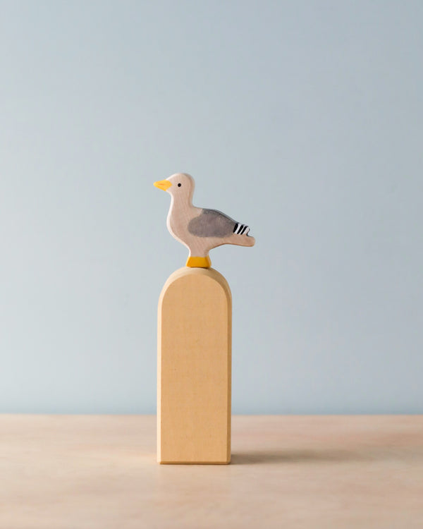 A handcrafted wooden bird figurine with a yellow beak and grey wings stands on top of a tall rectangular wooden block. Made in Europe from premium materials, this Holztiger Seagull is set against a light pastel blue background, while the block rests on a light brown surface.