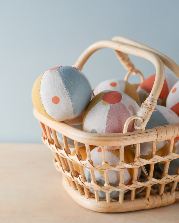 A wicker basket filled with colorful Maileg Rattle Balls sits on a light wooden surface. The balls, reminiscent of soft toy rattle balls, are patterned with pastel shades of blue, pink, yellow, and white, each adorned with polka dots. The playful accent against the plain light blue background adds charm.