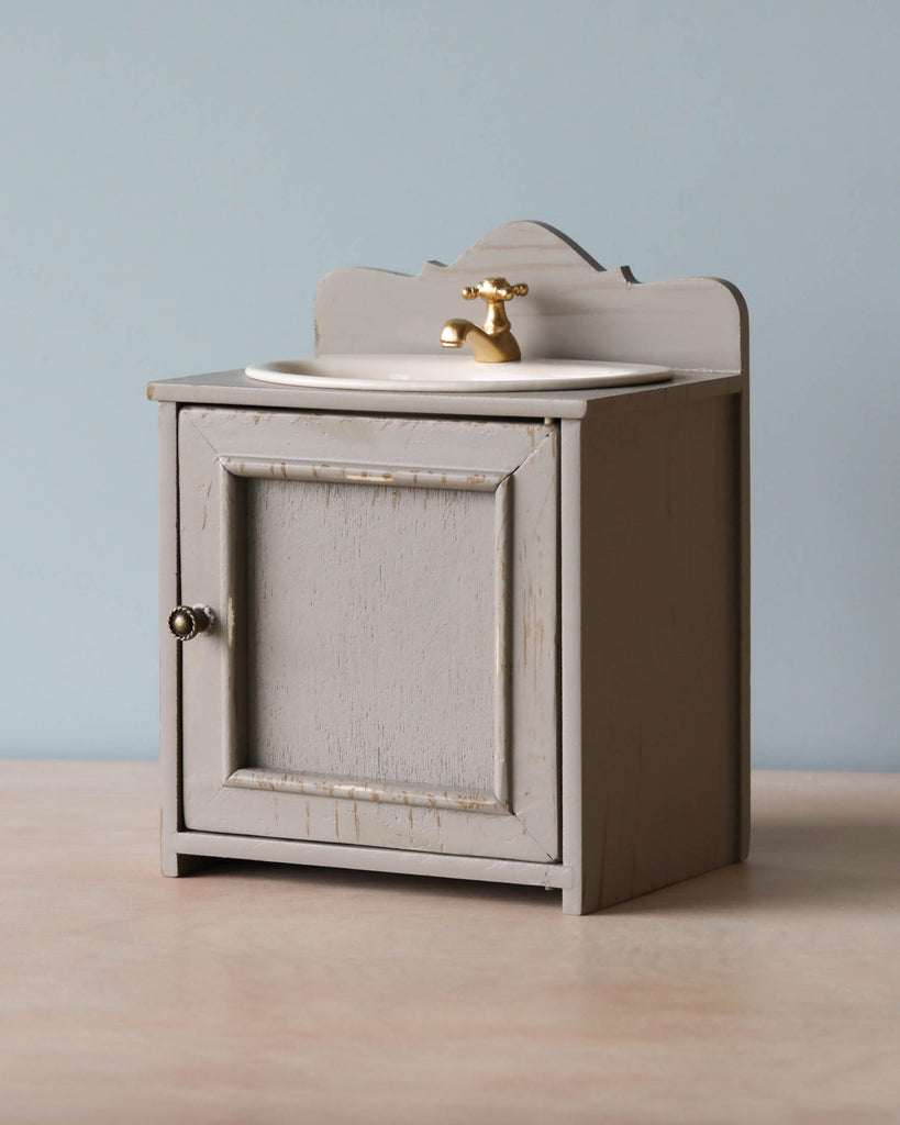 A small, vintage-style wooden nightstand painted in light beige, featuring a single cabinet door and a rounded top with an elegant Maileg | Miniature Sink fixture, against a pale blue background.