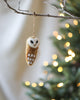 A Handmade Felt Owl Ornament, made in Nepal, hanging on a branch, with twinkling lights softly blurred in the background, creating a festive and cozy ambiance.