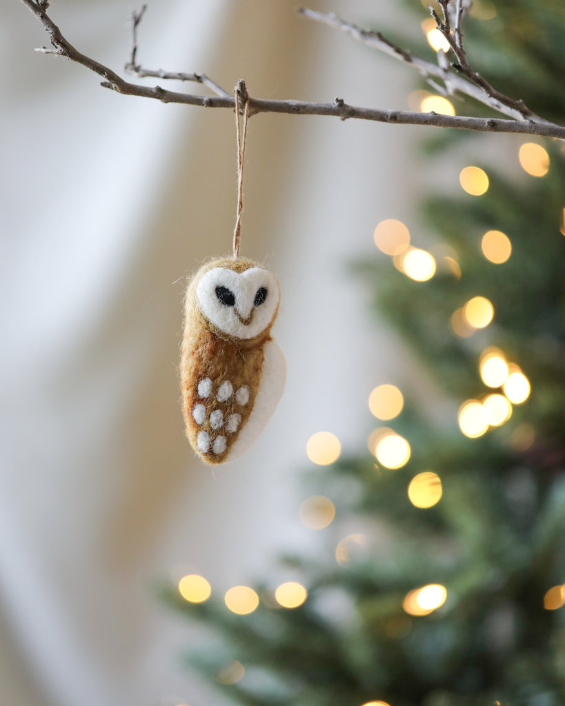 A Handmade Felt Owl Ornament, made in Nepal, hanging on a branch, with twinkling lights softly blurred in the background, creating a festive and cozy ambiance.