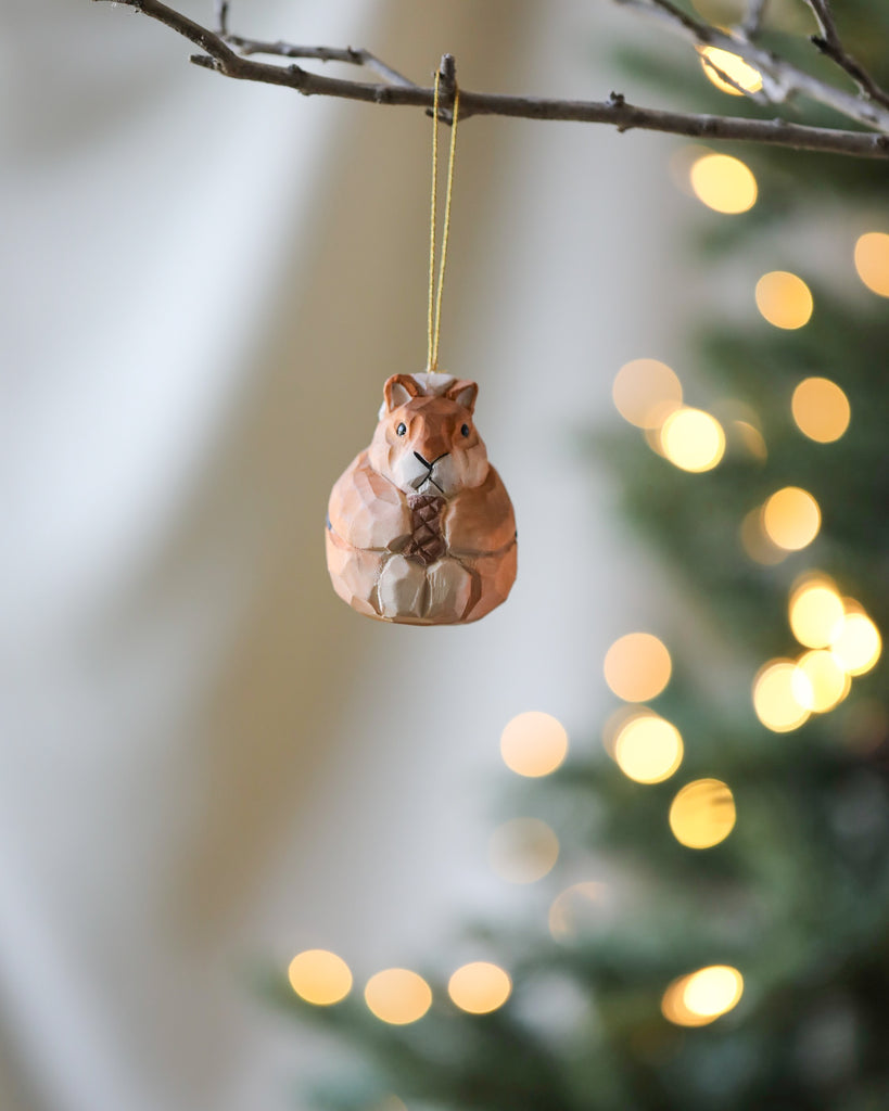 A decorative Wooden Ornament hangs from a tree branch, set against a softly blurred background with twinkling golden lights.