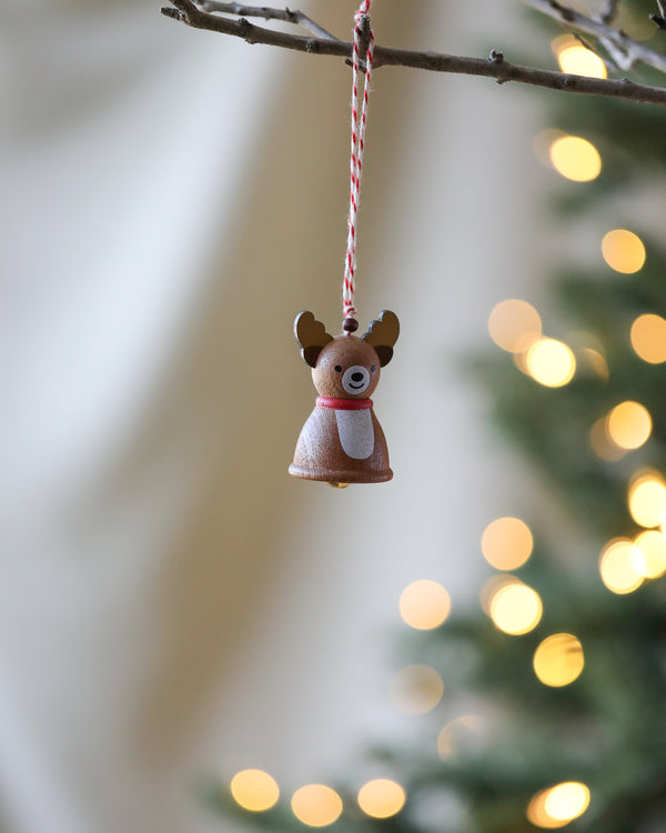 A small **Wooden Reindeer Ornament**, crafted from natural wood, hangs on a red and white string against a blurred background of a Christmas tree with golden lights.