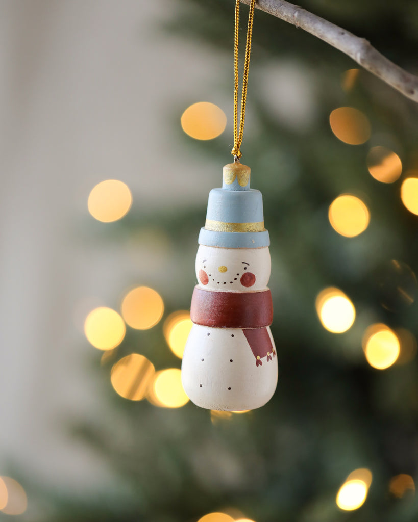 A Handmade Wooden Snowman Ornament, standing 3.5" tall, hangs from a branch against a backdrop of warm, glowing lights. The snowman has rosy cheeks, a blue and white hat, a red scarf, and black buttons down its front, creating a festive and cozy holiday atmosphere.