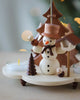 A Christian Ulbricht Snowman Pyramid, handmade in Germany, with a bird on its hand stands next to a pyramid candleholder and a miniature pine tree, set against a softly blurred background with festive lights.