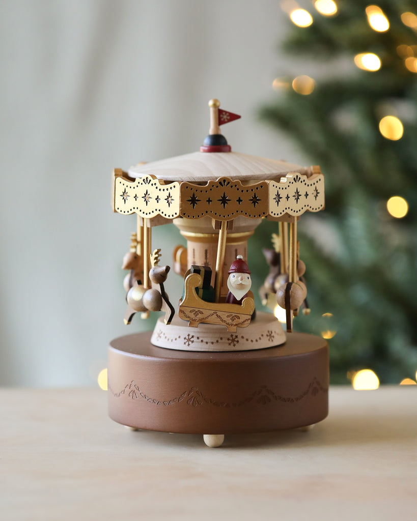 A festive, decorative Christmas Carousel Music Box on a table, featuring Santa in a sleigh and reindeer. This hand-cranked music box with classic design plays "Joy To The World" as it rotates. The carousel has intricate details, lit by warm, blurred background lights, with a Christmas tree partially visible to the right.