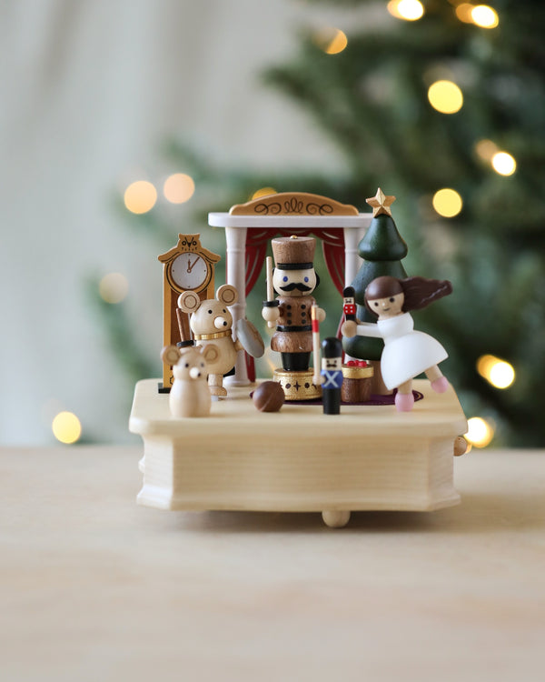 A wooden holiday display featuring Nutcracker Ballet Music Box figurines, a snowman, and a decorated tree on a Wooderful Life music box carousel, with a softly blurred Christmas tree in the background.