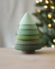 A Raduga Grez | Fir Tree Stacker with layers painted in varying shades of non-toxic green paint. The background shows a blurred, lit Christmas tree with warm white lights, creating a soft, cozy atmosphere. This piece embodies sustainability and is perfect for an eco-friendly holiday decor.