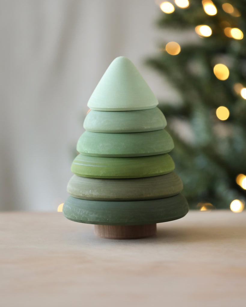 A Raduga Grez | Fir Tree Stacker with layers painted in varying shades of non-toxic green paint. The background shows a blurred, lit Christmas tree with warm white lights, creating a soft, cozy atmosphere. This piece embodies sustainability and is perfect for an eco-friendly holiday decor.