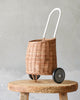 A unique Olli Ella Rattan Luggy, hand-woven from rattan, with a white handle, displayed on a rustic wooden stool against a textured gray background.