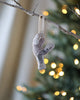 A handcrafted Coral & Tusk Ice Skater Bunny Ornament hangs from a branch, with a blurred background featuring twinkling Christmas tree lights. The ornament is detailed, emphasizing its unique stitching and design, resembling the shape of a bunny.