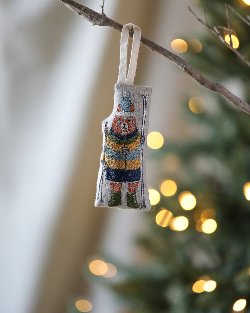 The Coral & Tusk Slopes Fox Ornament, featuring a fox in a striped parka and pom-pom cap holding skis, hangs from a branch with soft, blurred Christmas tree lights in the background.