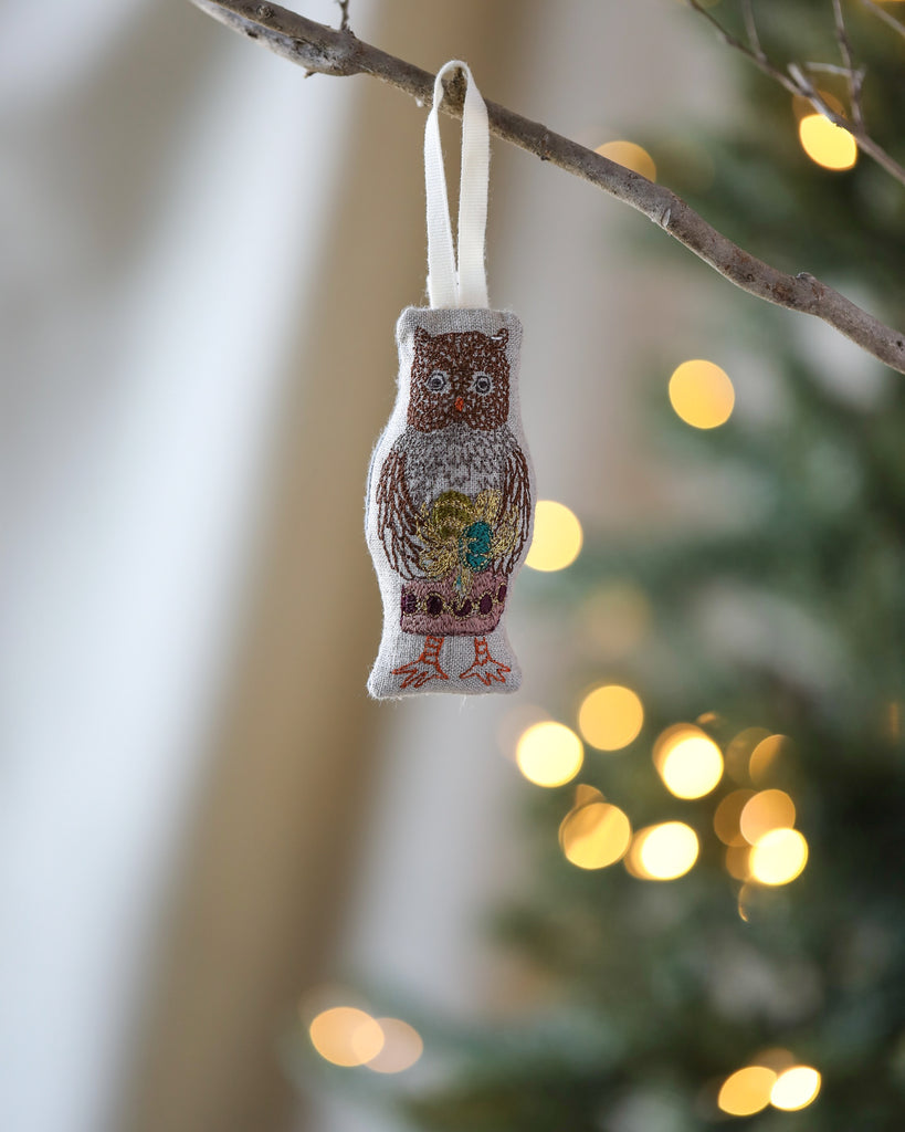 The Coral & Tusk Owl Ornament, with embroidered details, hangs from a branch, capturing the festive forest theme. In the background, warm white lights on a blurred Christmas tree add to the cozy holiday ambiance among other beautiful Christmas ornaments.