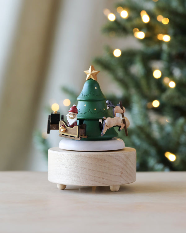 The Santa & Reindeer Music Box is a festive, hand-cranked music box featuring a miniature Christmas tree topped with a star, encircled by Santa and reindeer on a rotating platform. Made from sustainably sourced wood, it serves as the ideal decoration against the backdrop of a softly lit Christmas tree.