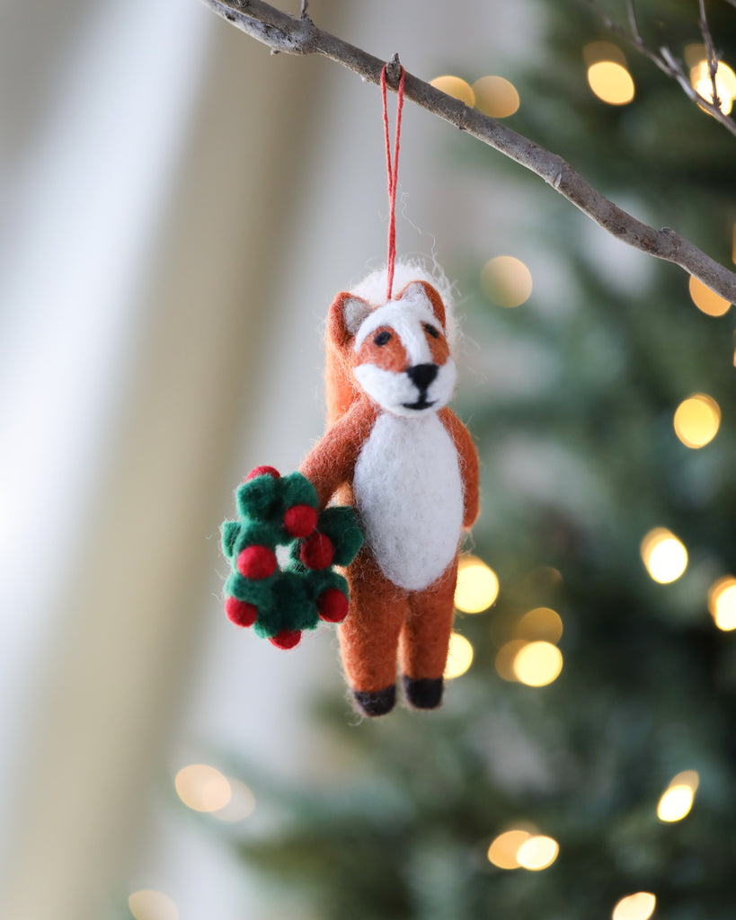 A Handmade Felt Fox Christmas Tree Ornament holding a handmade felt wreath, hanging from a Christmas tree with blurred lights in the background.