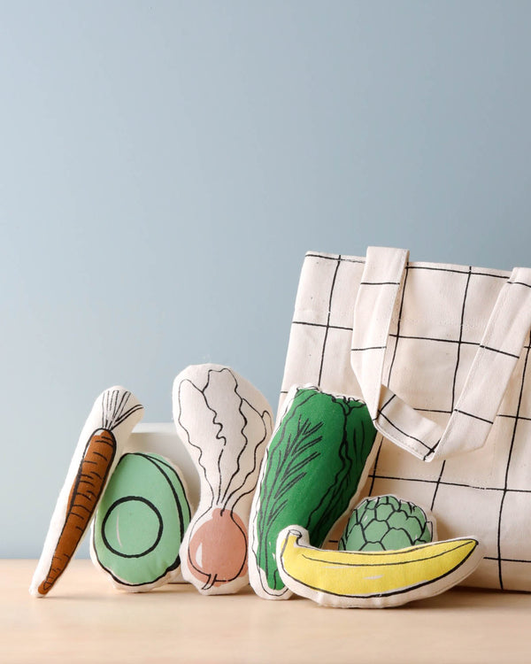 A collection of plush toy vegetables consisting of a carrot, radish, avocado, cabbage, and banana, alongside a Farmer’s Market Tote on a wooden table against a pale blue background.