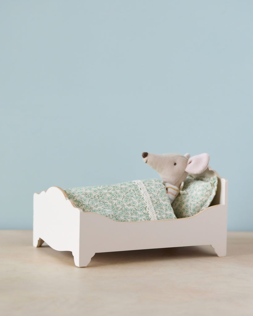 Sentence with product name: A Maileg Wooden Bed tucked into a small wooden bed with a green patterned bed linen, against a plain blue backdrop.