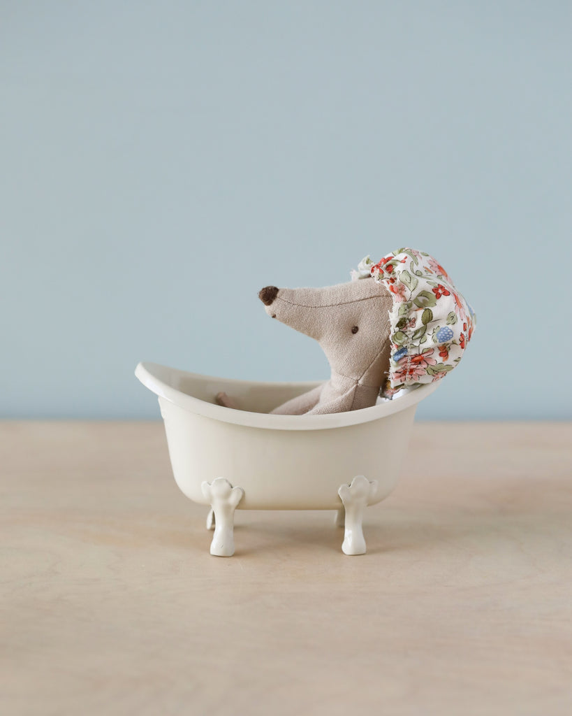 A small stuffed mouse, affectionately dubbed the Wellness Mouse, wears a floral patterned shower cap as it relaxes in a Maileg Bathtub (Miniature) on a wooden surface against a light blue background.