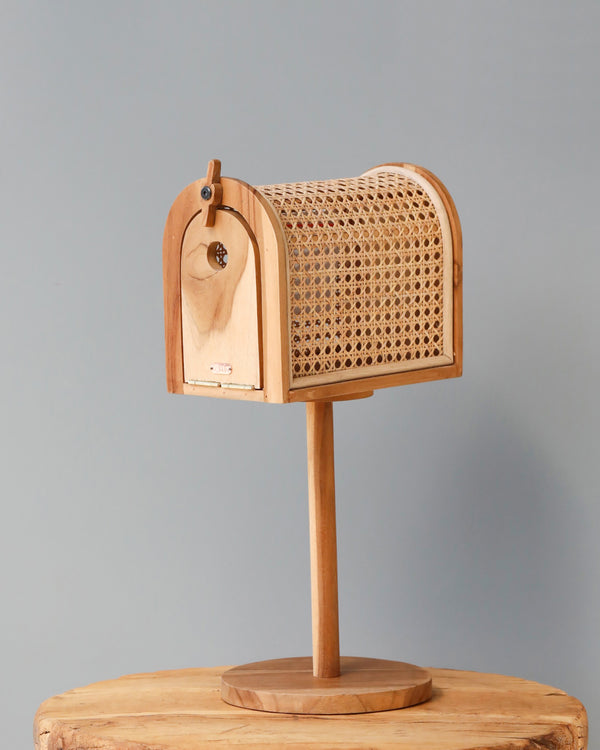 Poppie Rattan Mailbox on a stand, featuring a curved rattan webbing door and a small, round brass lock, set against a plain gray background.