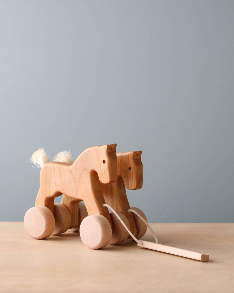 A Galloping Horses Pull Toy with wheels and a pull string, featuring a simple carved design and fluffy white tail, set against a soft grey background.