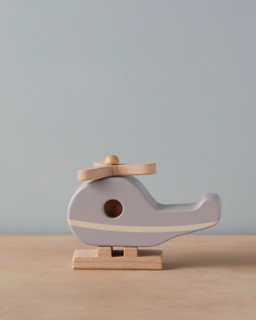 A minimalist Wooden Helicopter with a soft gray body and responsibly sourced beech wood rotors and skids, displayed against a pale blue background.
