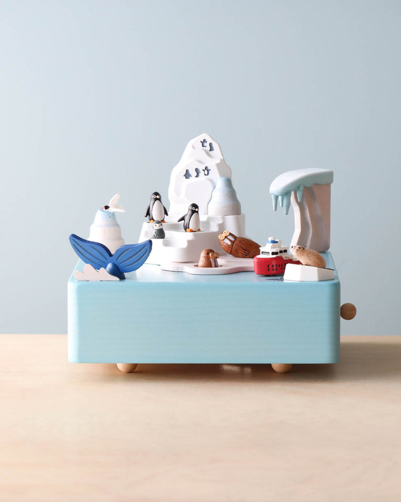A whimsical hand cranked Wooden Antarctica Music Box featuring nautical elements like a lighthouse, seagulls, a sailboat, and a whale's tail, set against a pale blue and cream backdrop.