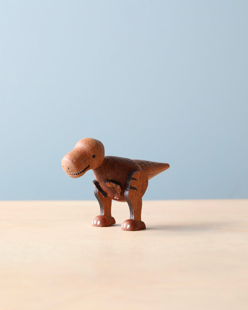 A Wooden Dinosaurs toy, resembling a tyrannosaurus rex, stands on a table against a pale blue background. The dinosaur is crafted with visible wood grain and joints, making it an ideal aspiring paleontologist's toy.