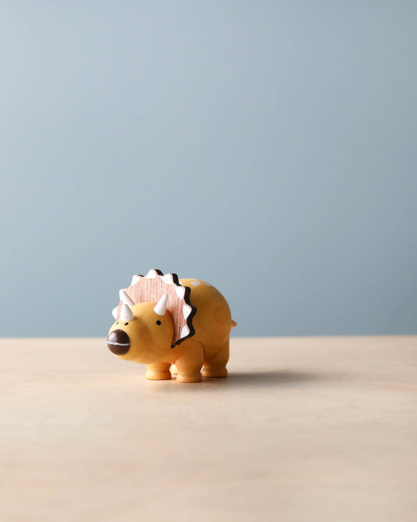 A wooden dinosaur figurine with hand-painted details stands on a wooden surface against a soft blue background. The dinosaur has its spikes styled into a playful black and white pattern, perfect as a