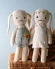 Two Cuddle + Kind Bunny dolls standing on a wicker basket. One wears gray overalls with a carrot pocket detail, and the other, in a white dress, has a floral headband.
