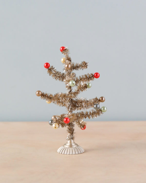 The Maileg Christmas Tree, Small - Gold with tinsel branches and adorned with red, gold, beige, and green round ornaments stands as a festive centerpiece on a small white base against a light gray background. Nearby, a gingerbread house adds to the holiday charm.