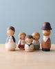 A collection of five Handmade Peg Doll - Mom figurines representing a family, displayed on a table against a soft blue background. Each figurine has unique, simplistic facial features and detailed clothing.