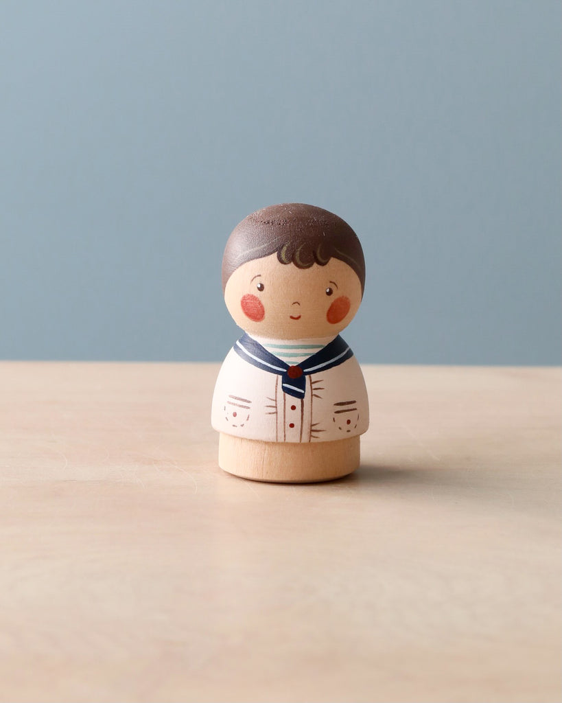 A Handmade Peg Doll - Boy with dark hair, wearing a white outfit with blue accents, standing on a light brown surface against a pale blue background.