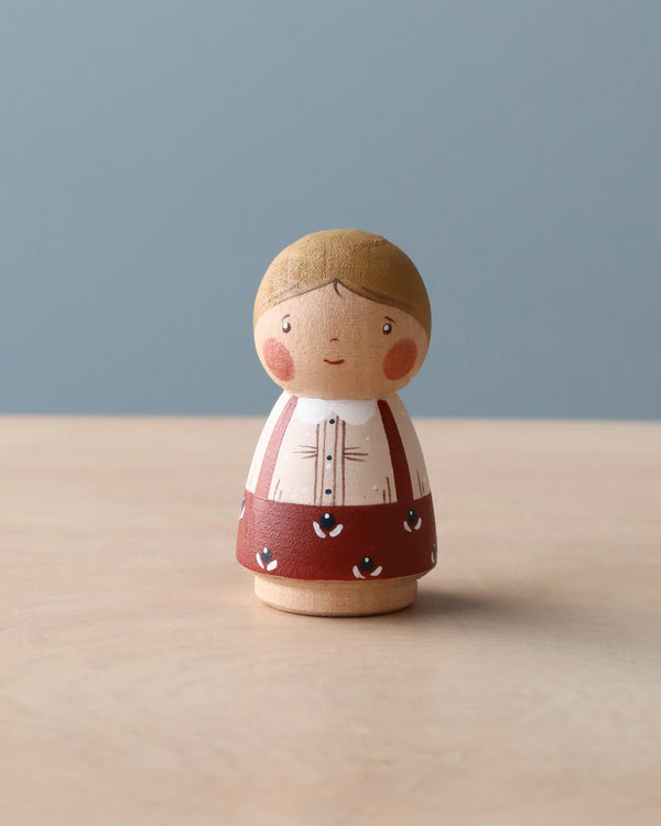 A hand-painted wooden peg doll designed to resemble a small girl with braided hair, standing on a wooden surface against a soft blue background.