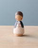 A handmade wooden Mom peg doll stands on a wooden table against a pale blue background. The doll depicts a person with a light brown hairstyle, wearing a white dress adorned with blue and red accents.