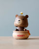 A Wooden Honey Bear Bobblehead with a friendly face sitting in a honey pot that has "honey" written on it, with a tiny bee perched on top of the bear’s head, against