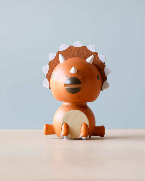 A Wooden Triceratops Bobblehead with a round body, painted in an orange-brown color, featuring white spikes along its back and a cute, simplified face. It sits against a plain light blue
