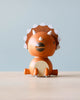 A Wooden Triceratops Bobblehead with a round body, painted in an orange-brown color, featuring white spikes along its back and a cute, simplified face. It sits against a plain light blue