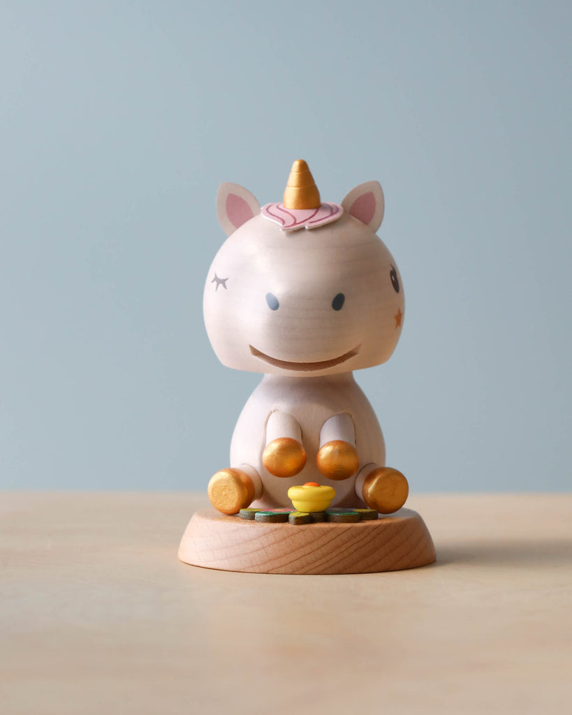 A Wooden Unicorn Bobblehead toy with a pastel color palette, featuring a pink horn and ears, seated with a xylophone in front on a light blue background