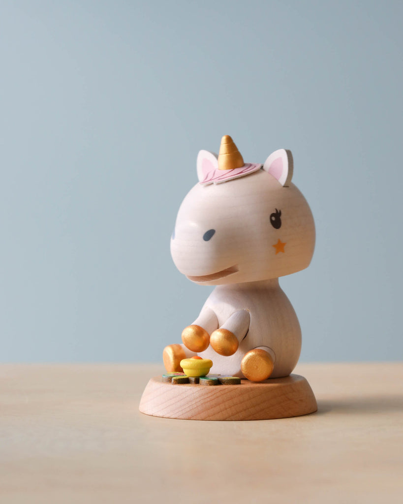 The Wooden Unicorn Bobblehead with a pink and gold horn, winking eye, and star detail stands on a wooden base, holding tiny, colorful mushrooms against a soft blue background.