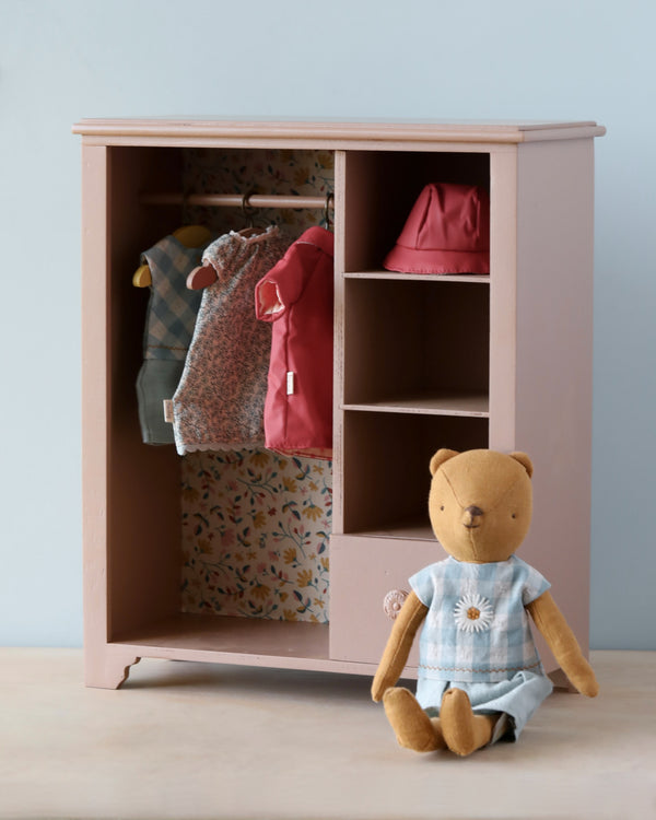 The Maileg Large Wooden Wardrobe is a charming piece adorned with floral wallpaper inside, featuring doll-sized clothes on hangers and pink accessories on shelves. Next to the wardrobe sits a stuffed bear in a blue dress. This delightful children's toy storage is set on a light wooden surface against a pale blue background.