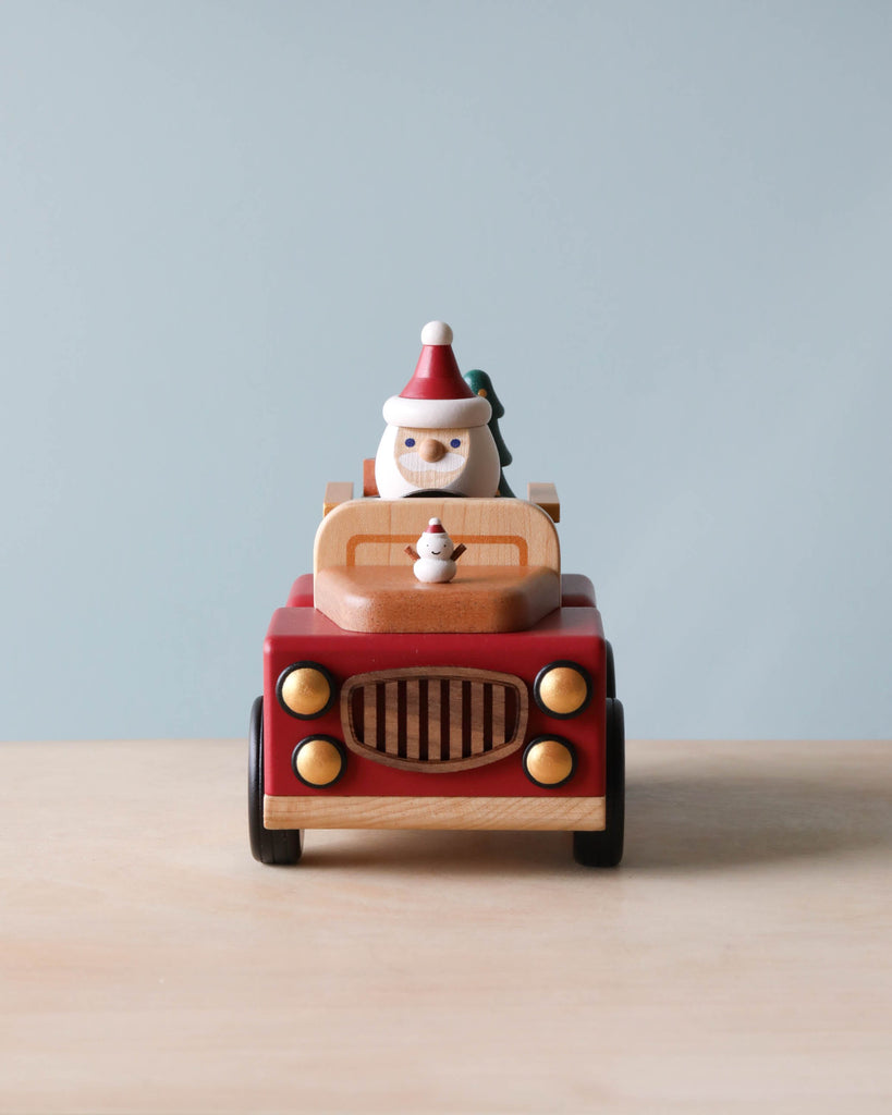 A Santa-mobile Music Box, crafted from premium hardwoods, in red with a yellow grille, carries a small figurine wearing a pointy hat driving against a plain blue background. A tiny snowman sits