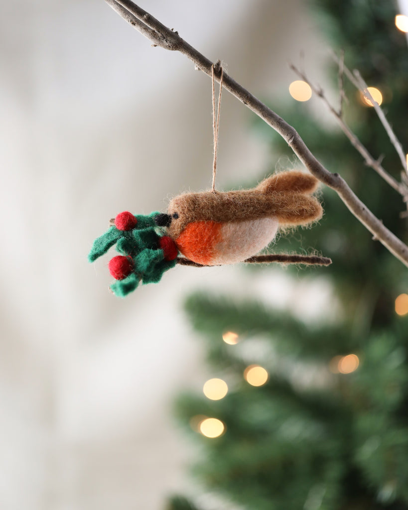 A Handmade Felt Robin on a Holly Branch Ornament with a red and green scarf holds a bundle of mistletoe, hanging from a branch with a blurred Christmas tree in the background.