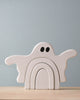 A Handmade Wooden Ghost Stacker, shaped like a ghost with outstretched arms and black eyes, handcrafted from poplar hardwood and finished with non-toxic wood dye, consists of three interlocking pieces. It is placed on a light wood surface against a light gray background.