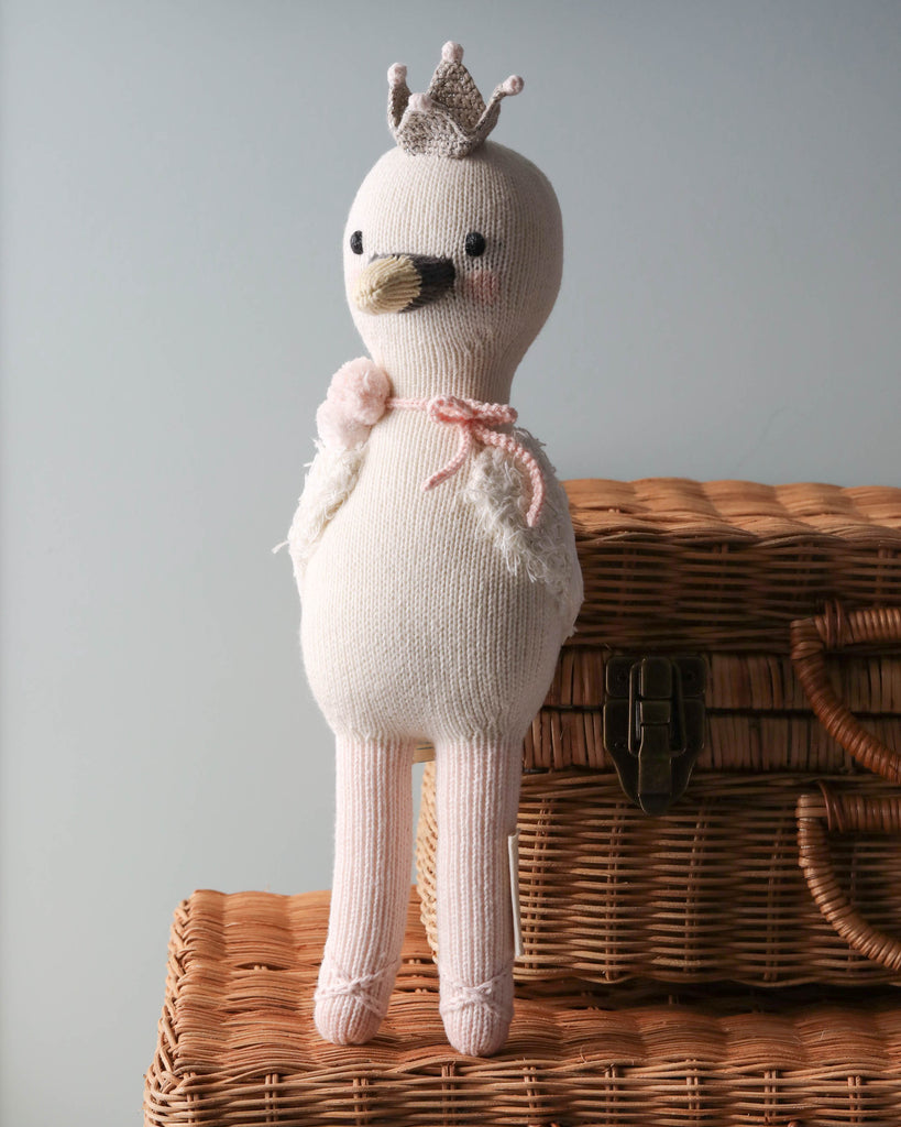 A Cuddle + Kind Harlow The Swan resembling a bird with a crown, sitting on a wicker basket. The doll is adorned with a small pink ribbon and has a serene expression.