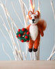 A Handmade Felt Fox Christmas Tree Ornament holding a green and red wreath hangs amid white painted branches against a soft blue background.