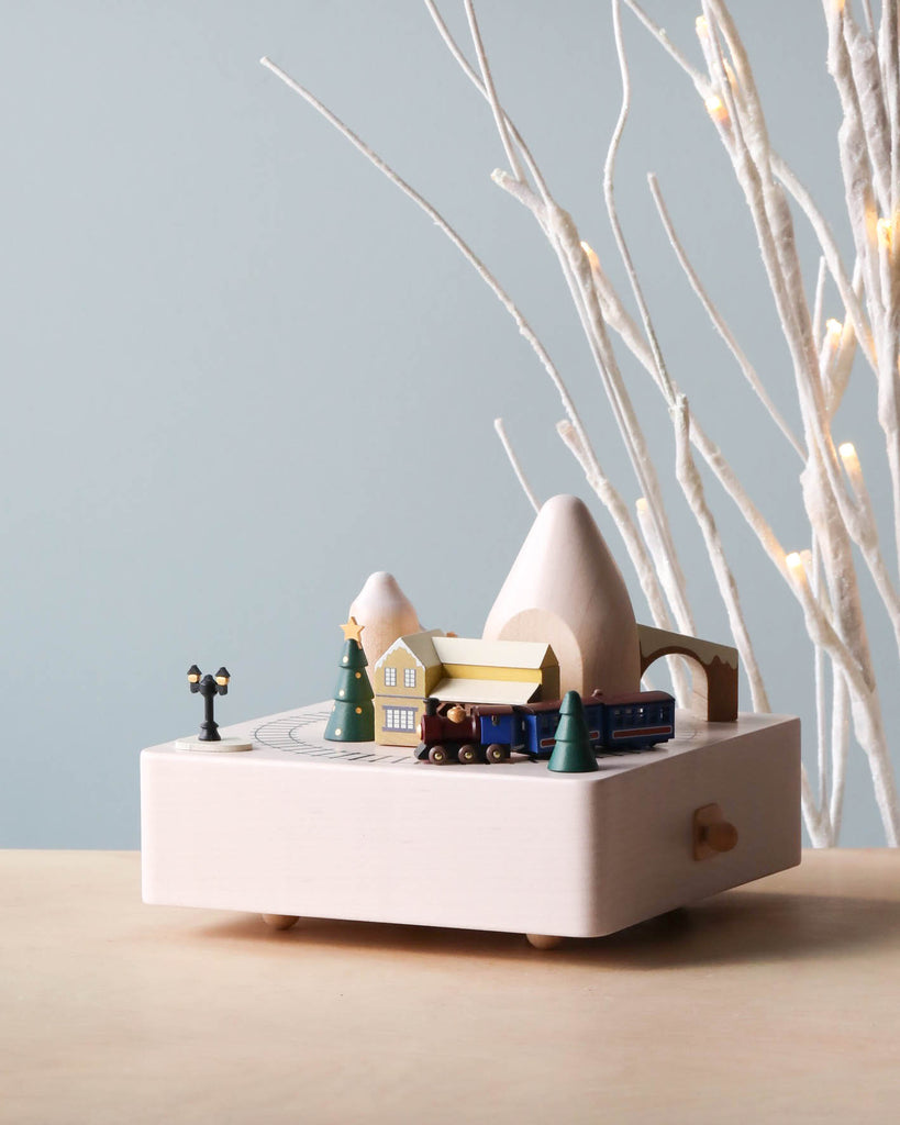 A minimalist wooden Christmas Train Music Box featuring tiny models of a mountain, house, and trees, with a figurine of a person, set against a soft blue background with white branches.