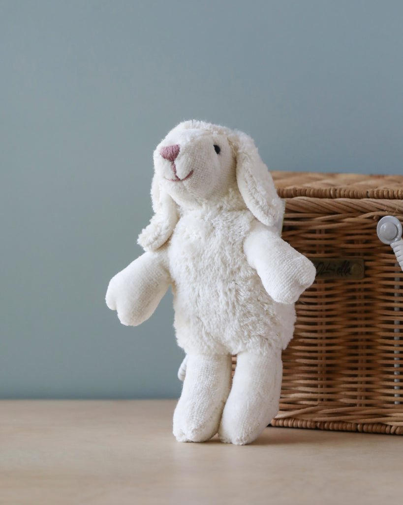 A Senger Naturwelt Stuffed Animal - Baby Sheep, handmade in Germany and crafted from organically grown cotton, with floppy ears is displayed standing on a wooden floor beside a wicker basket, set against a pale blue wall.
